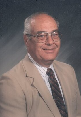 A school picture of Gurney during his time as principal of the Brown School. He is wearing a tan suit with glasses and is in front of a blue-gray background.
